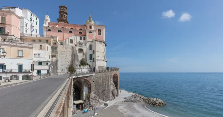Veduta di Atrani con le case e una chiesa affacciate sulla strada costiera, gli archi e la spiaggia
