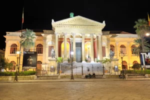 La facciata illuminata del Teatro Massimo di Palermo di notte, con colonne, scalinata e statue ai lati.