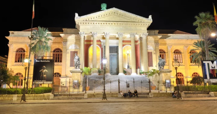 La facciata illuminata del Teatro Massimo di Palermo di notte, con colonne, scalinata e statue ai lati.